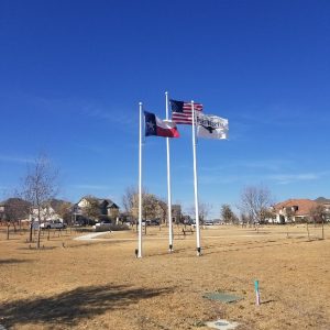Three flags flying on poles against a clear blue sky in an open field.