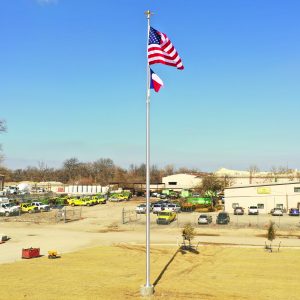 American and Texas flags fly on a tall flagpole in a rural area with buildings and vehicles.
