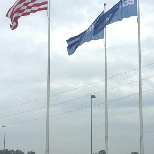 American and Greek flags waving on tall poles against a cloudy sky.