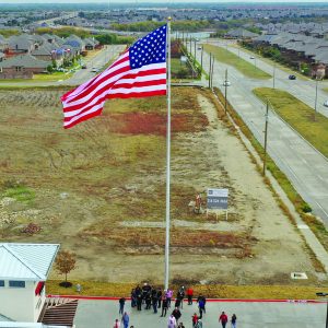 Large American flag flies high over an open area with people gathered below.